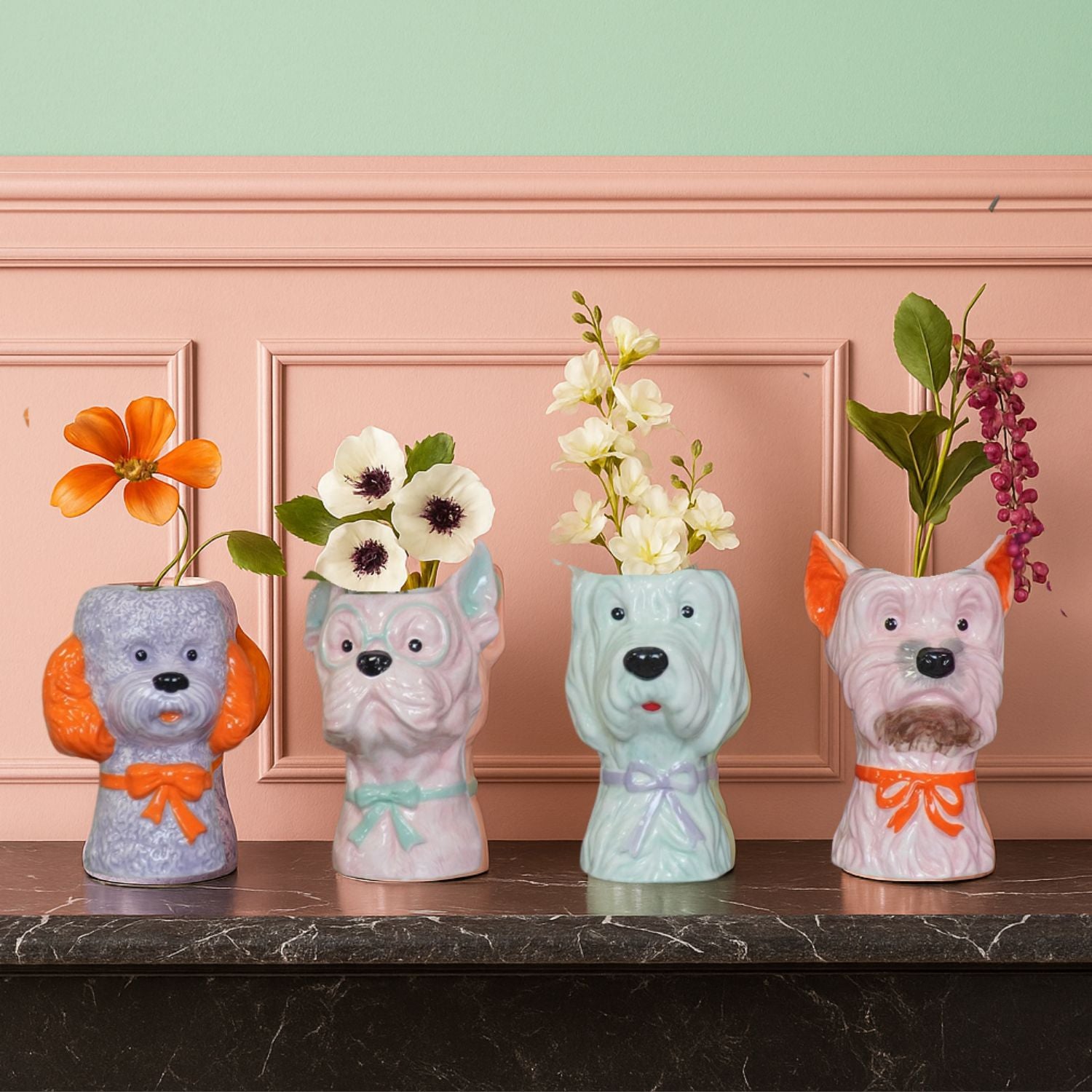 Four colourful dog-shaped vases with flowers, displayed on a marble surface against a pink panelled wall with a mint green upper section.