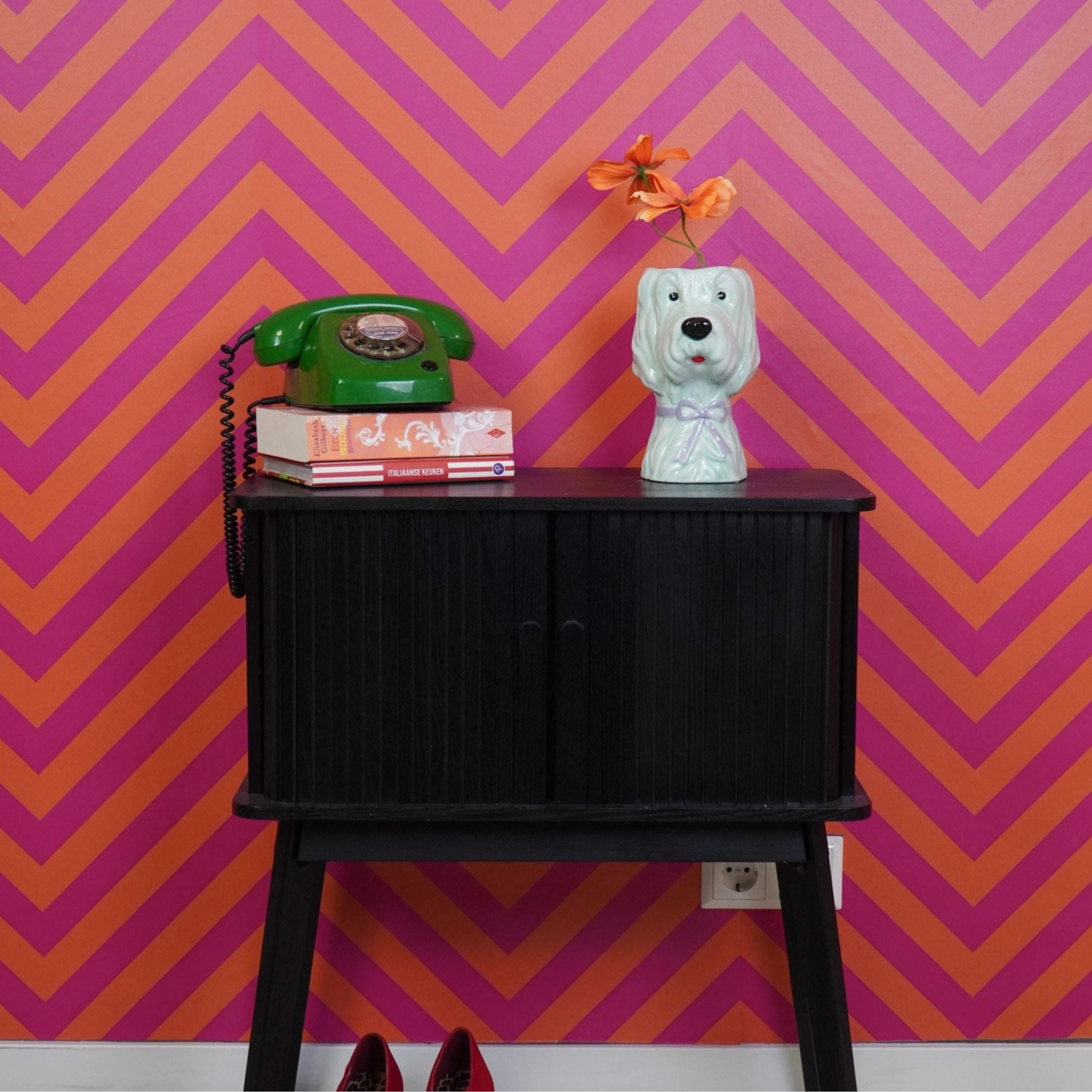 Mint green ceramic dog vase with orange flowers displayed on a black cabinet with a retro green telephone, set against a bold pink and orange chevron patterned wall.