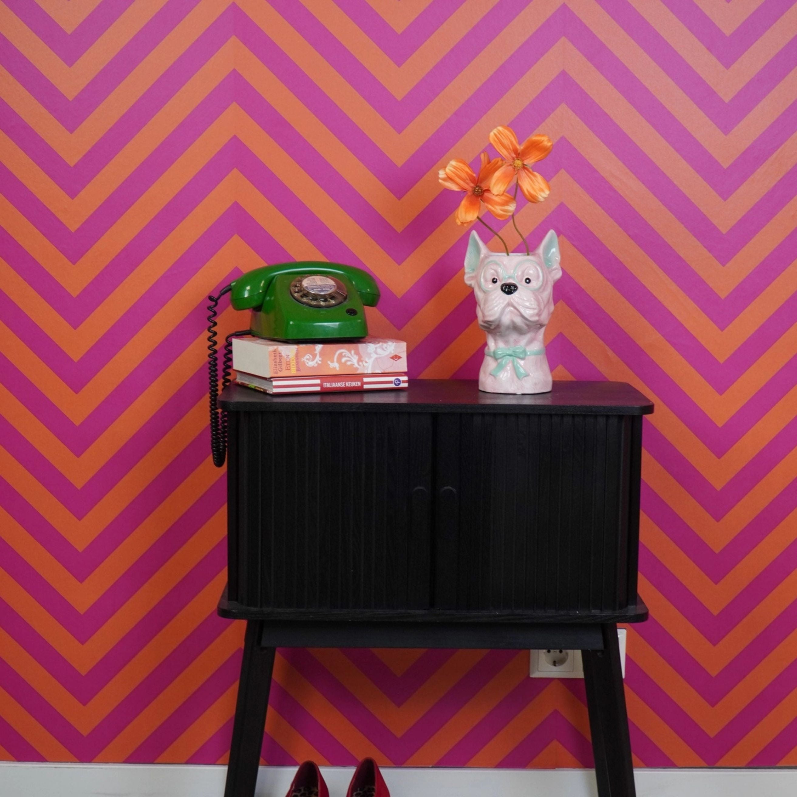Pink dog-shaped vase with mint green glasses and bow tie displayed on a black cabinet beside a green retro phone and books, against a vibrant pink and orange chevron wall