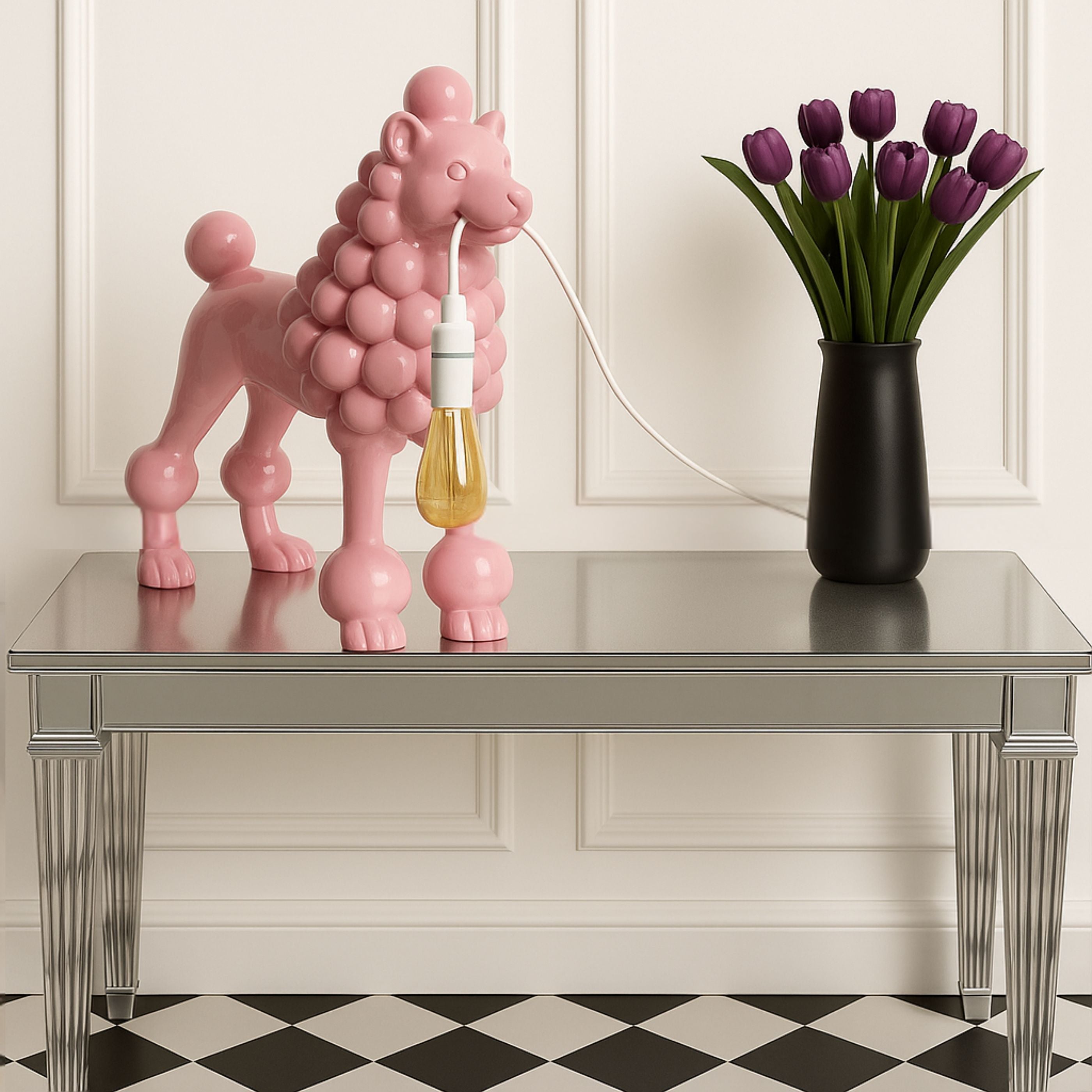 Pink poodle lamp with light bulb displayed on a silver console table beside a black vase of purple tulips, against a white panelled wall with black-and-white tiled floor