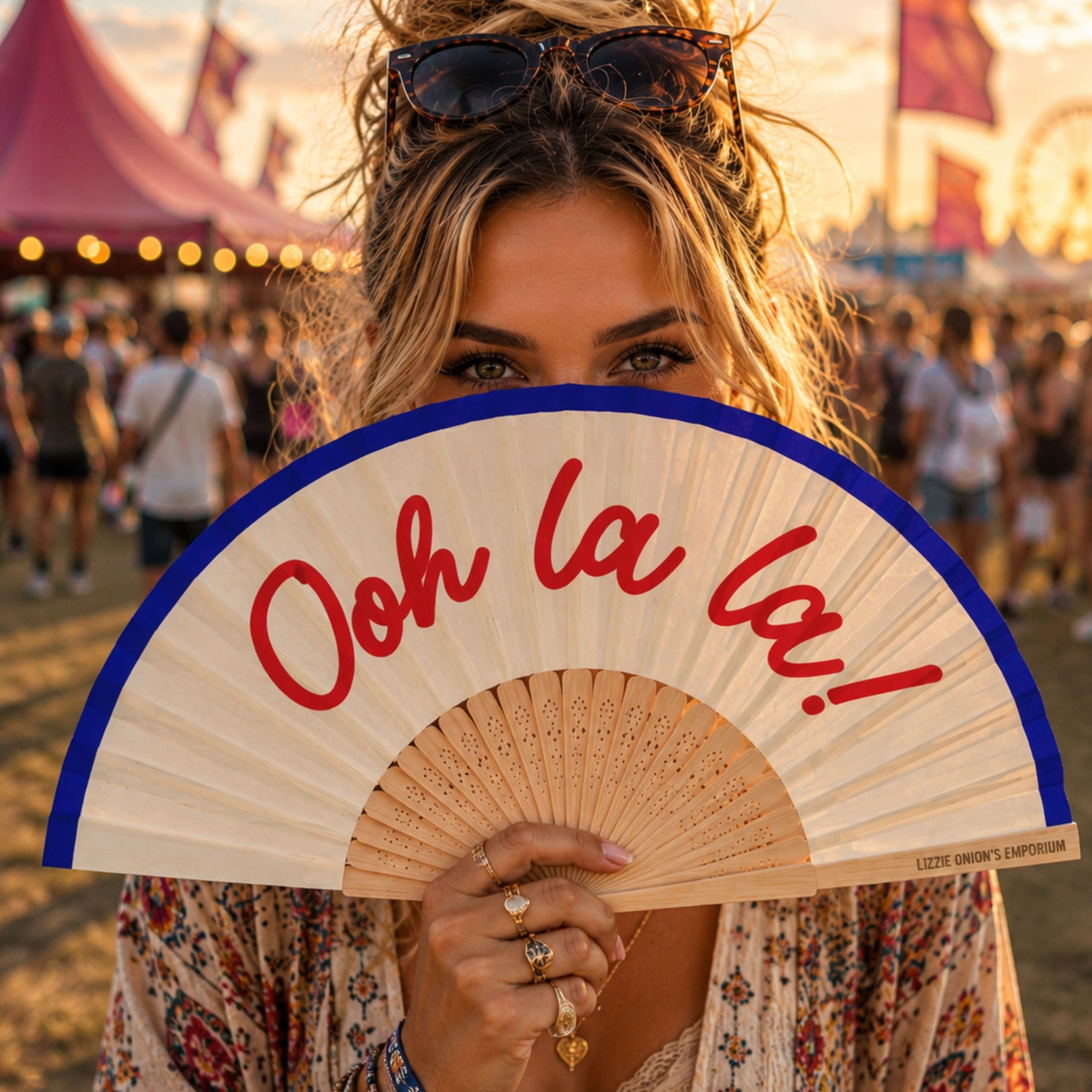 Woman holding Ooh La La folding hand fan at a summer festival, with only her eyes visible above the fan against a colourful festival backdrop.