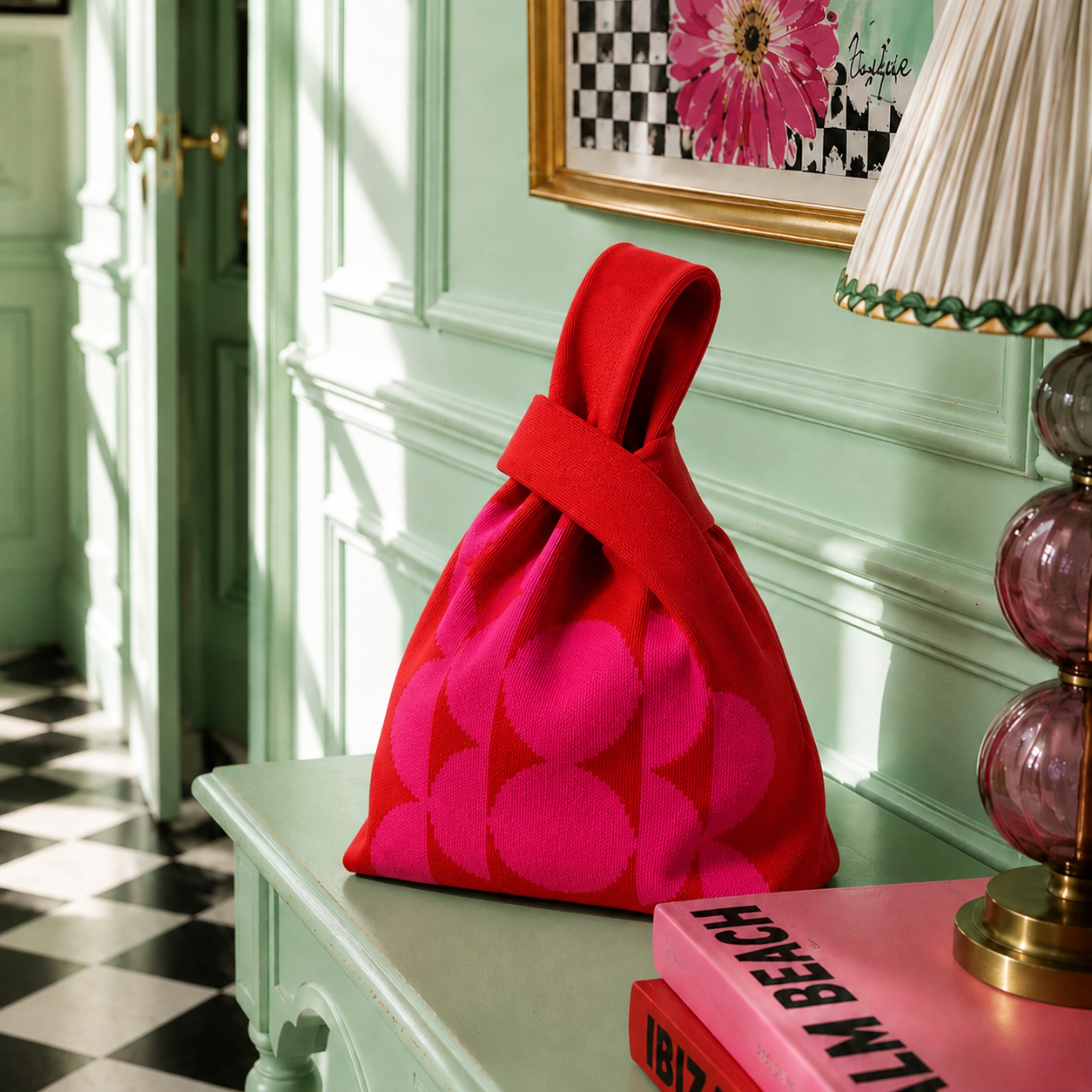 Bright red and hot pink knitted tote bag with a bold geometric pattern of oversized circles and semi-circles, styled on a mint green console table in a colourful maximalist interior. Warm natural sunlight streams through the space, highlighting the soft knitted texture, sculptural silhouette and vibrant colours, with black and white tiled flooring and playful décor creating a distinctive contemporary setting.