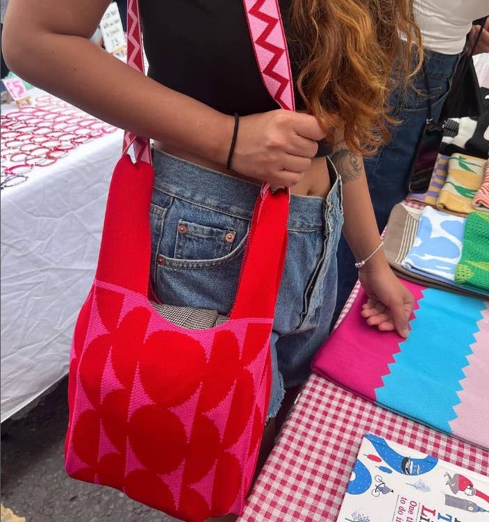 Bright red and hot pink knitted tote bag with a bold geometric pattern of oversized circles and semi-circles, worn over the shoulder at an outdoor market. The lightweight knitted fabric, spacious design and wide shoulder straps are shown in everyday use, highlighting the bag's comfortable fit, soft texture and vibrant contemporary style.