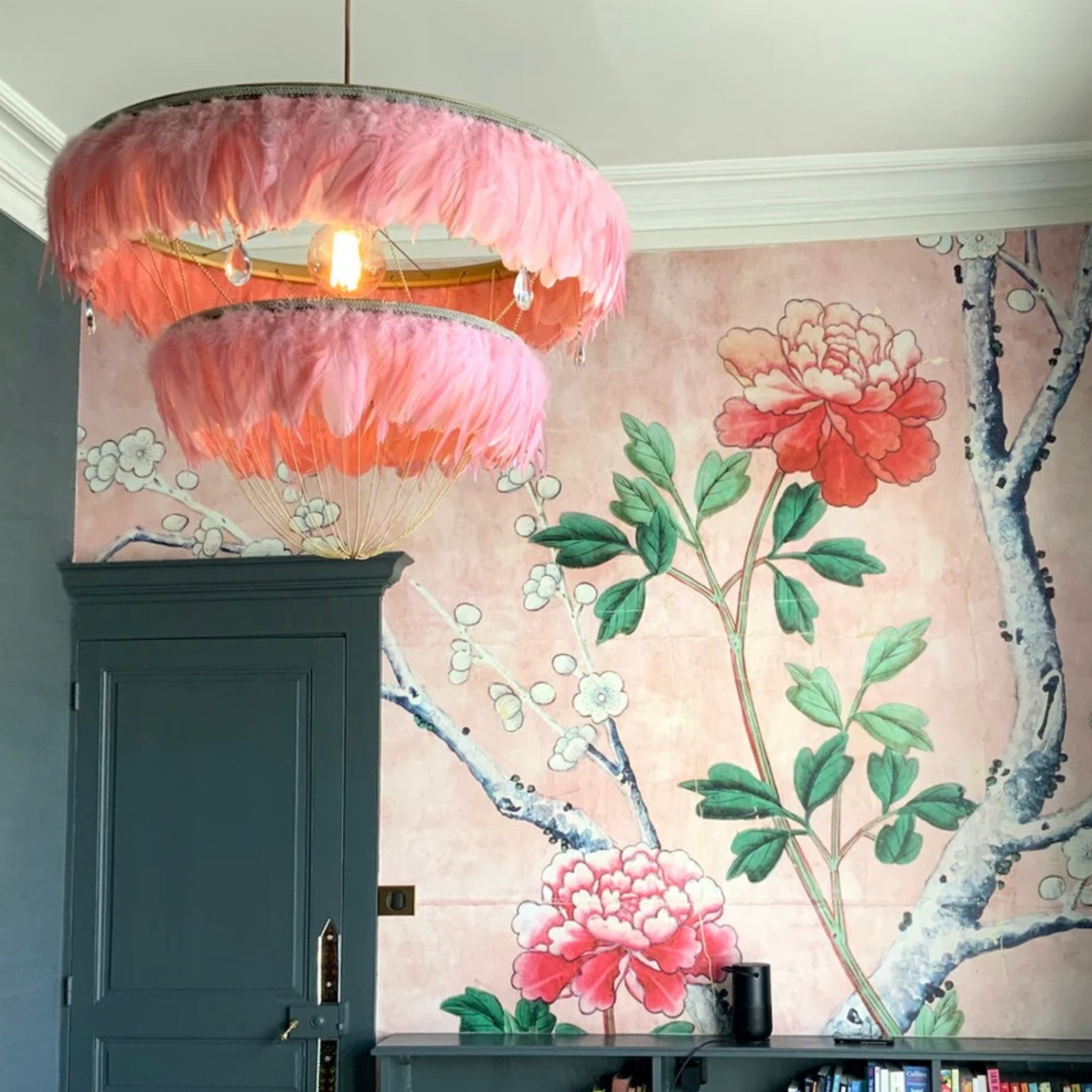 Flamingo pink two-tier Bertie feather light shade with crystal drops, hanging in front of a floral wallpapered wall and dark teal door.