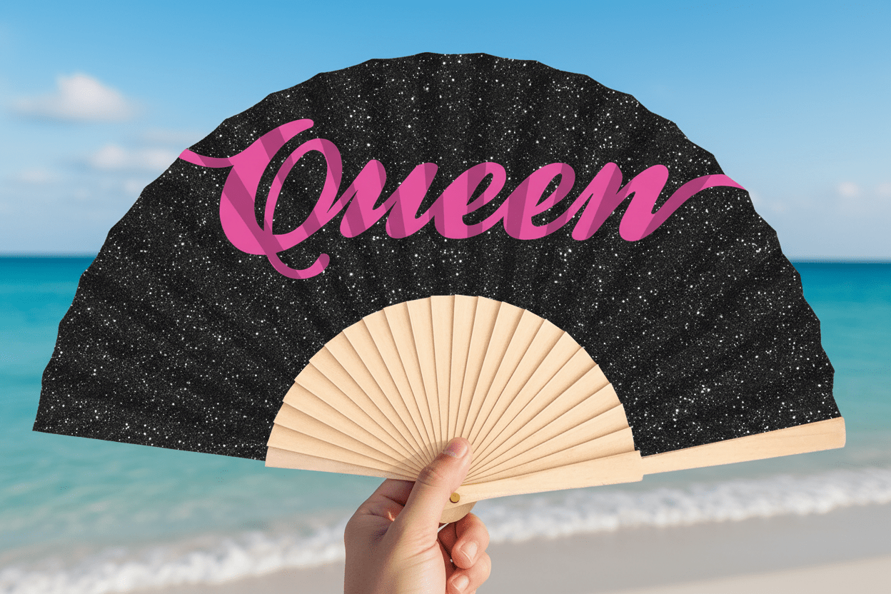 Hand holding a black glitter 'Queen' folding hand fan with pink slogan lettering on a tropical beach with turquoise sea in the background.