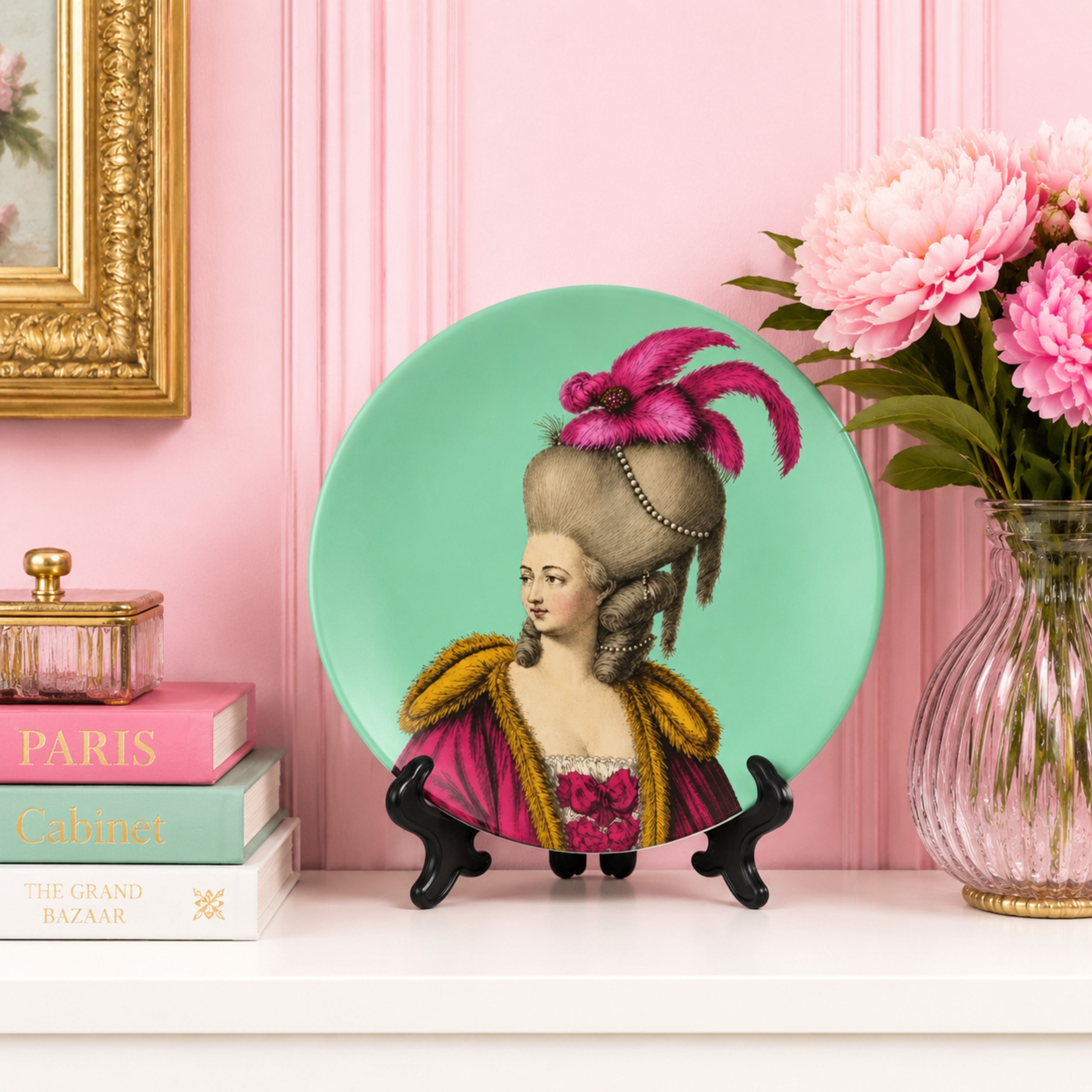 Lady Seraphine Featherstone decorative ceramic plate displayed on a black stand beside pastel books and pink flowers against a blush pink panelled wall in elegant maximalist style.