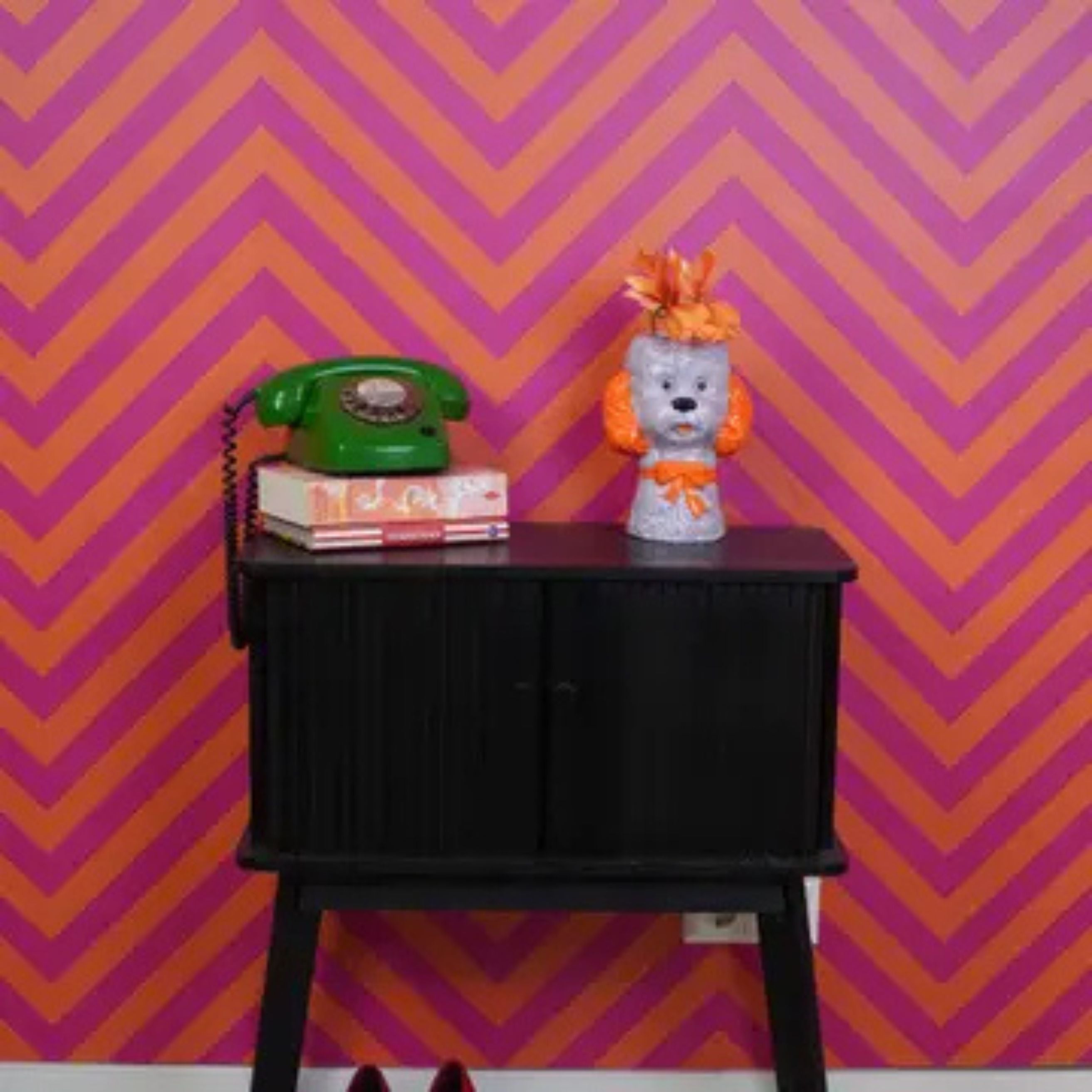 Lilac and orange dog-shaped ceramic vase with flowers styled on a black cabinet beside a green retro phone, against a pink and orange chevron wall.