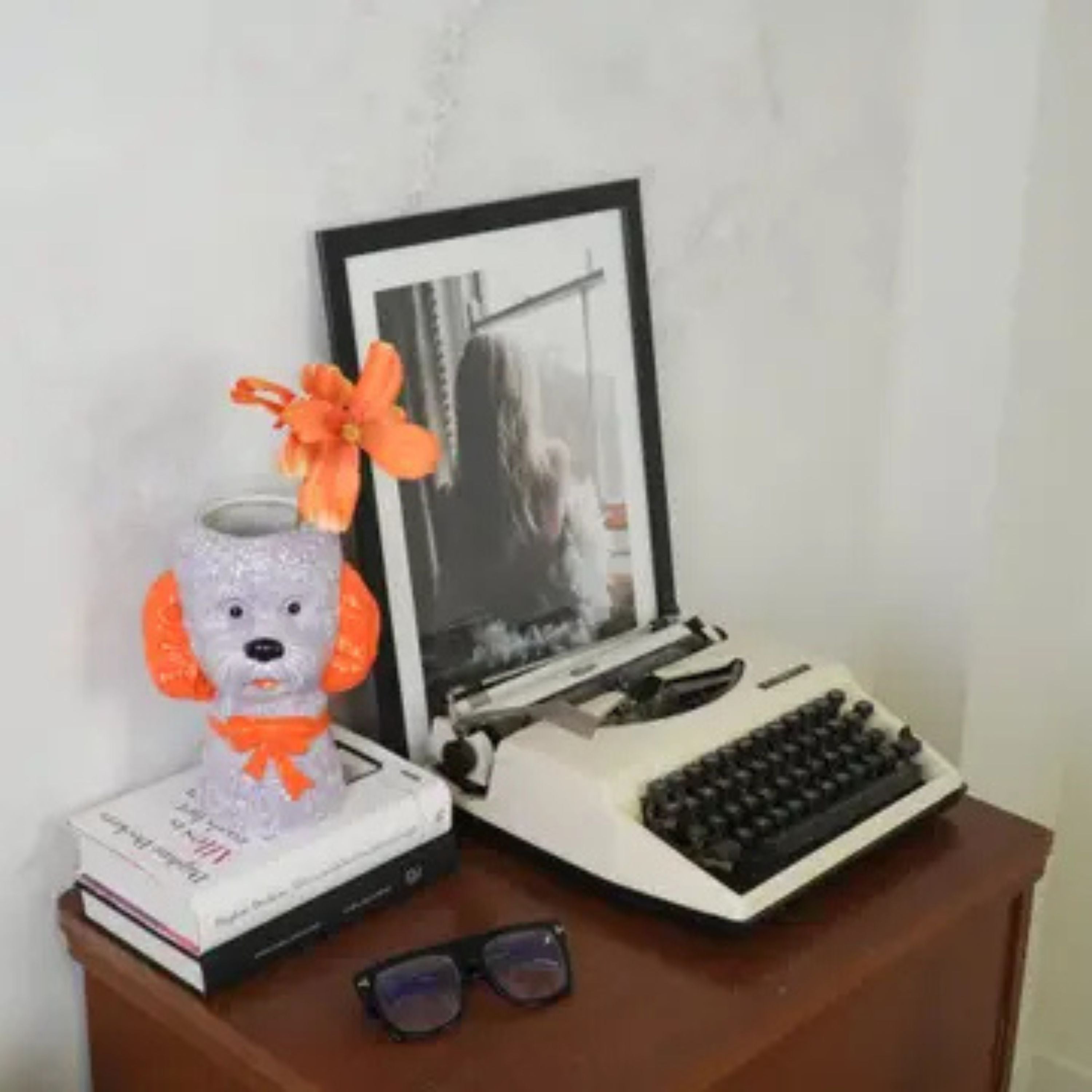 Lilac and orange dog-shaped ceramic vase with flowers styled on stacked books beside a vintage typewriter and framed photo on a wooden table.