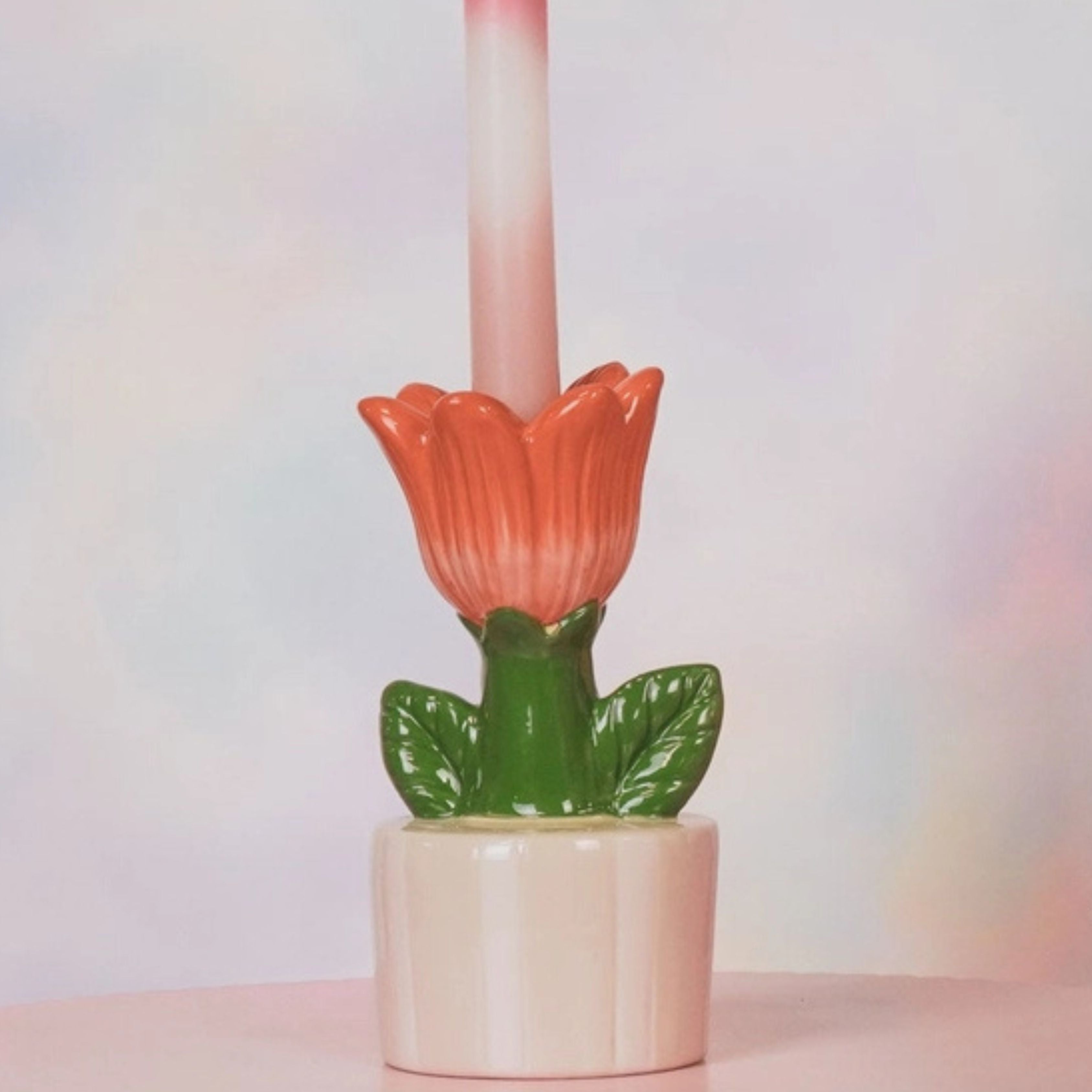 Orange tulip-shaped stoneware candle holder with green leaves holding a pink taper candle, shown against a soft pastel background.