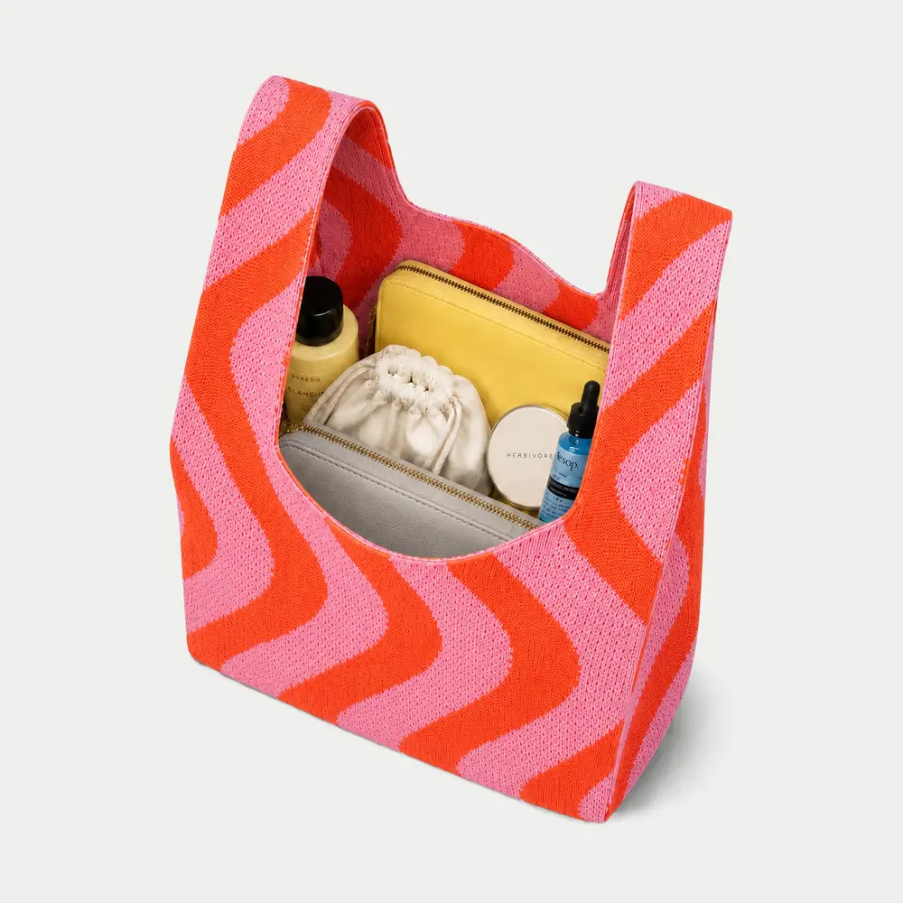 Sunset Swirl Knitted Bag filled with everyday essentials, showcasing the spacious interior. Lightweight knitted tote featuring a bold retro-inspired coral orange and candy pink wave pattern.