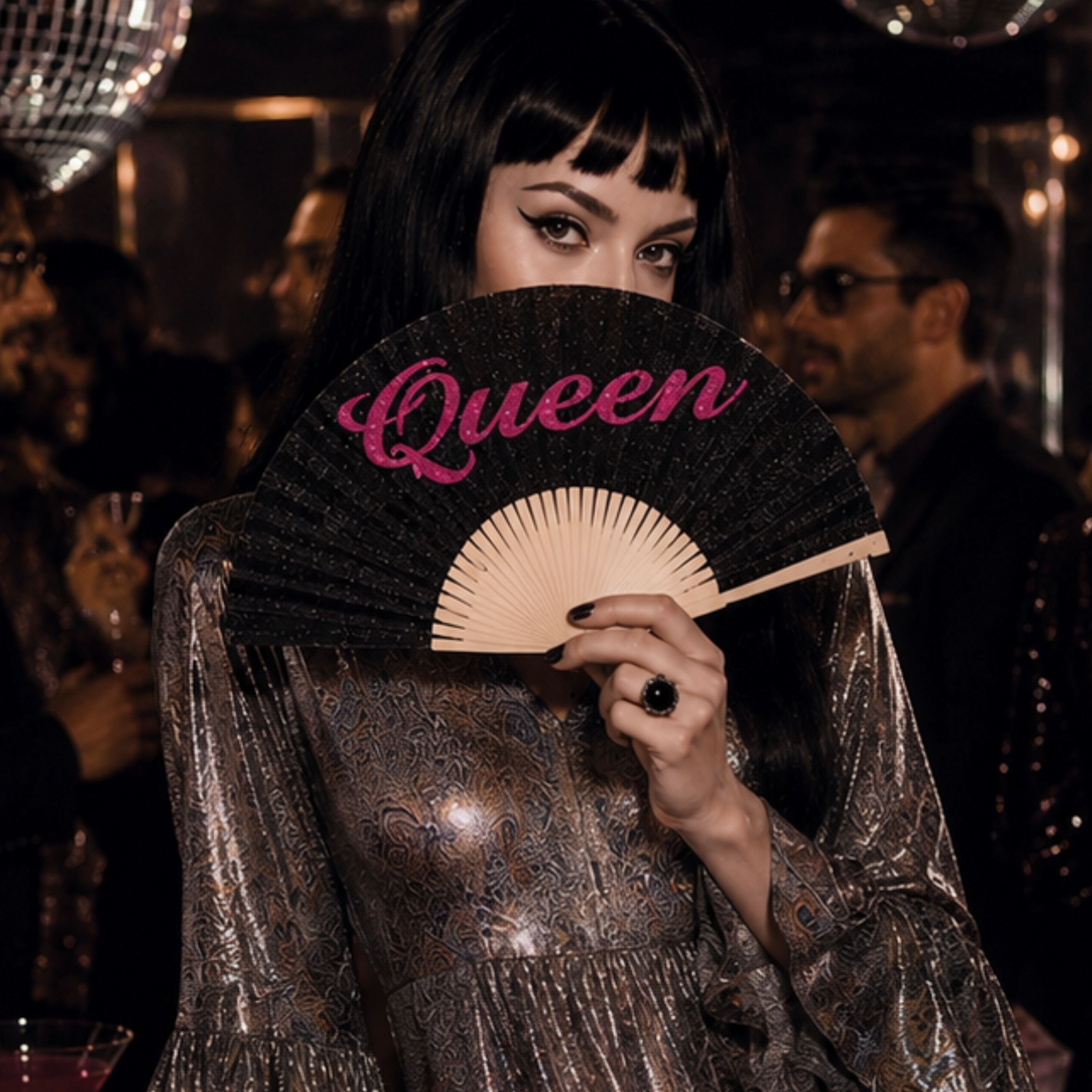 Woman holding a black glitter 'Queen' folding hand fan at a glamorous disco-inspired party, wearing a metallic ruffle dress with mirrored disco balls in the background.